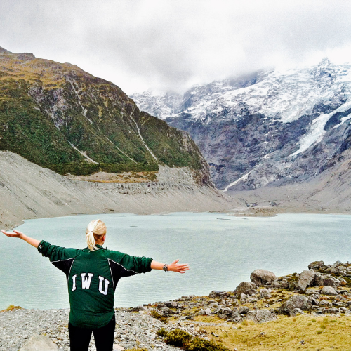 Student wearing IWU sweater extends arms in front of a mountain landscape