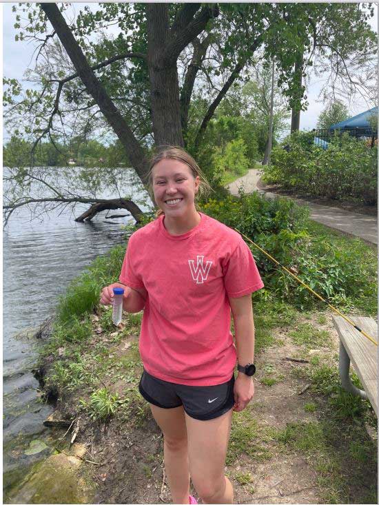 Amanda Wilson smiles to camera while standing next to a body of water and holding a vial.