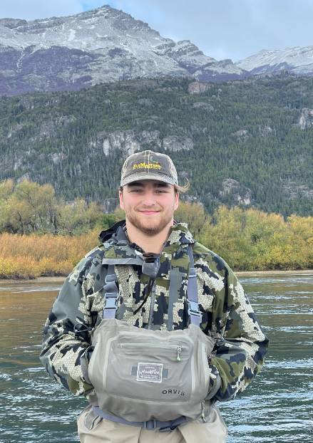 Male college student wearing hat and overalls, with large body of water, trees, and mountains in the background