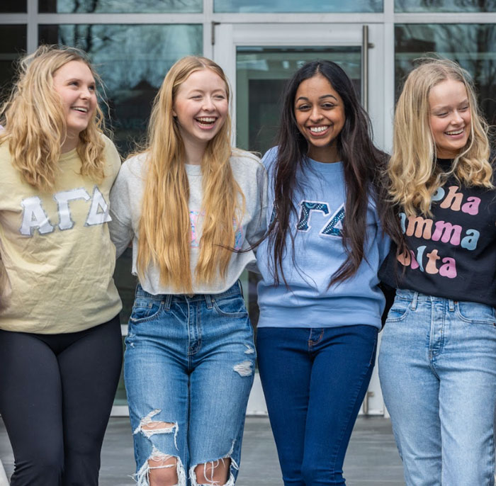 Four Alpha Gamma Delta sorority members stand in front of State Farm Hall, smiling and wearing Alpha Gamma Delta t-shirts
