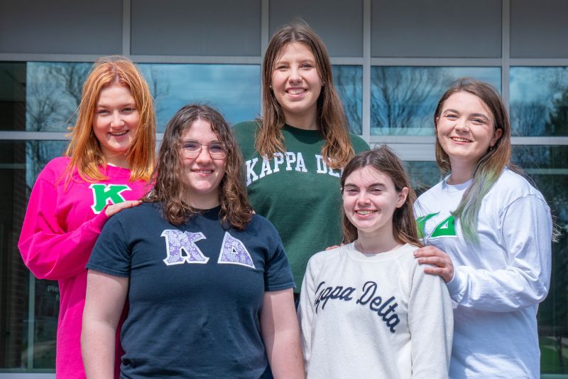 Five Kappa Delta sorority members stand in front of State Farm Hall smiling and wearing Kappa Delta letters and t-shirts