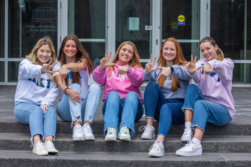 Five Kappa Kappa Gamma sorority members sit on the steps of State Farm Hall and throw their hand sign while smiling