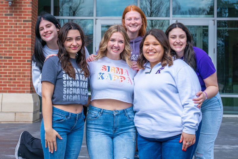 Six members of the Sigma Kappa sorority stand in front of State Farm Hall smiling and wearing Sigma Kappa t-shirts