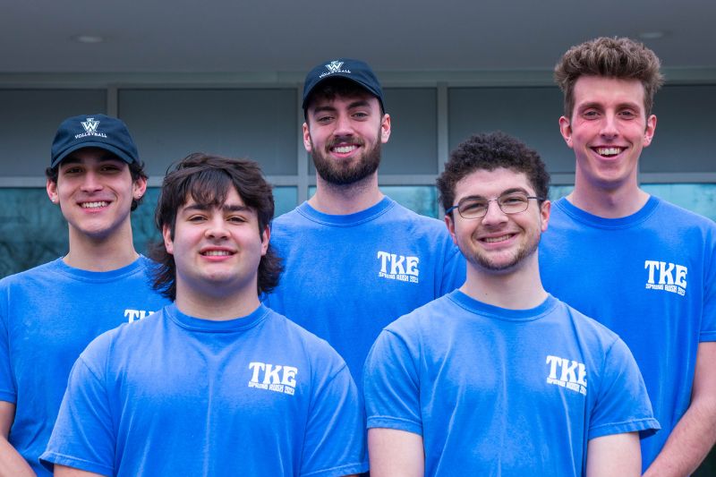 Five members of the Tau Kappa Epsilon fraternity stand in front of State Farm Hall smiling and wearing TKE shirts