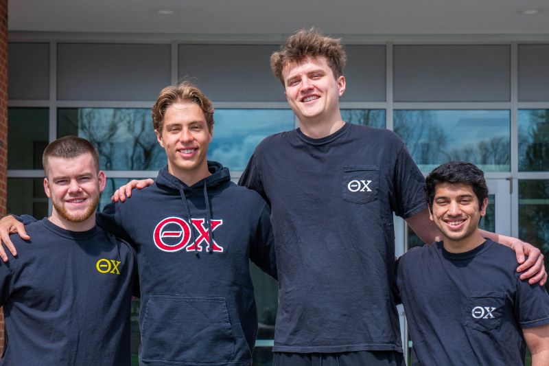 Four members of the Theta Chi fraternity stand in front of State Farm Hall smiling and wearing Theta Chi t-shirts