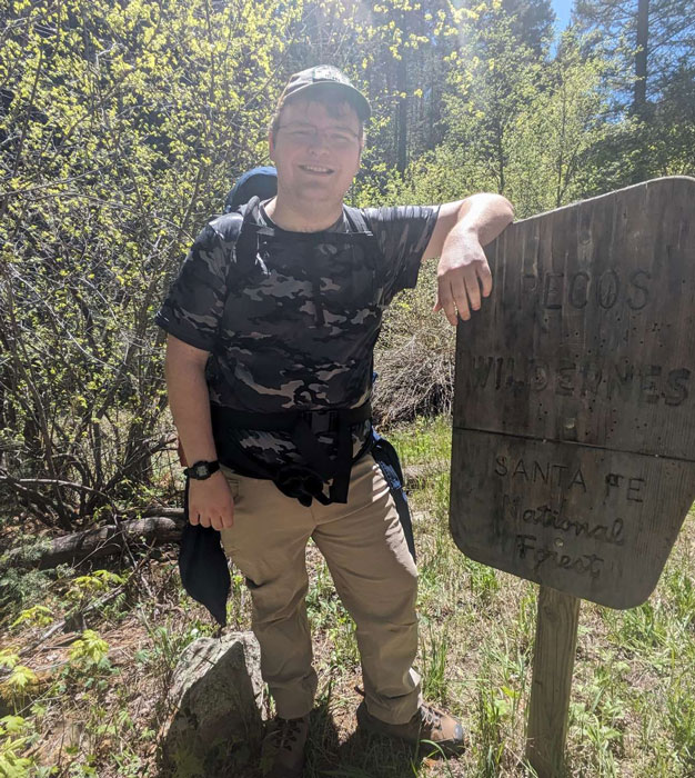 Male student wearing a backpack and smiling next to a hikinig trail