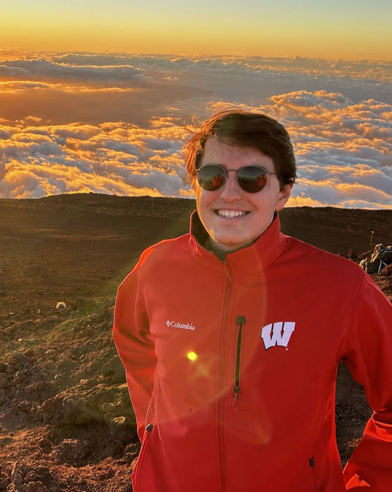 Male college student wearing sunglasses and smiling in front of a mountain view at sunset