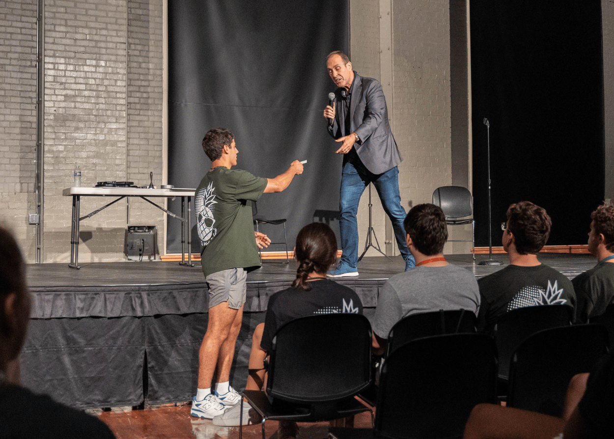 Chris Carter stands on stage in front of students, reaching out to accept a playing card selected by a student