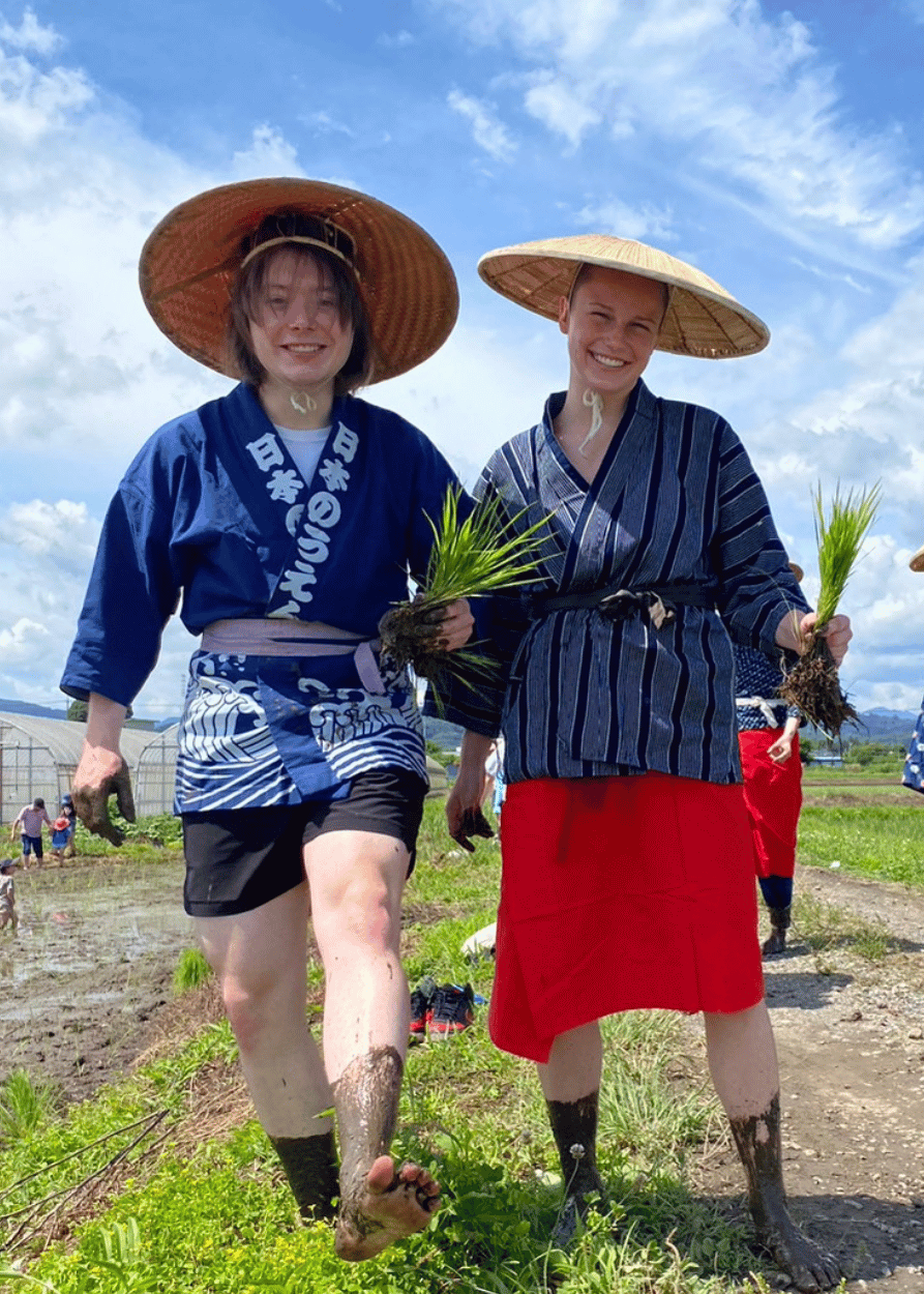 Two IWU students stand barefoot next to a rice field in Japan wearing traditional rice farming clothes