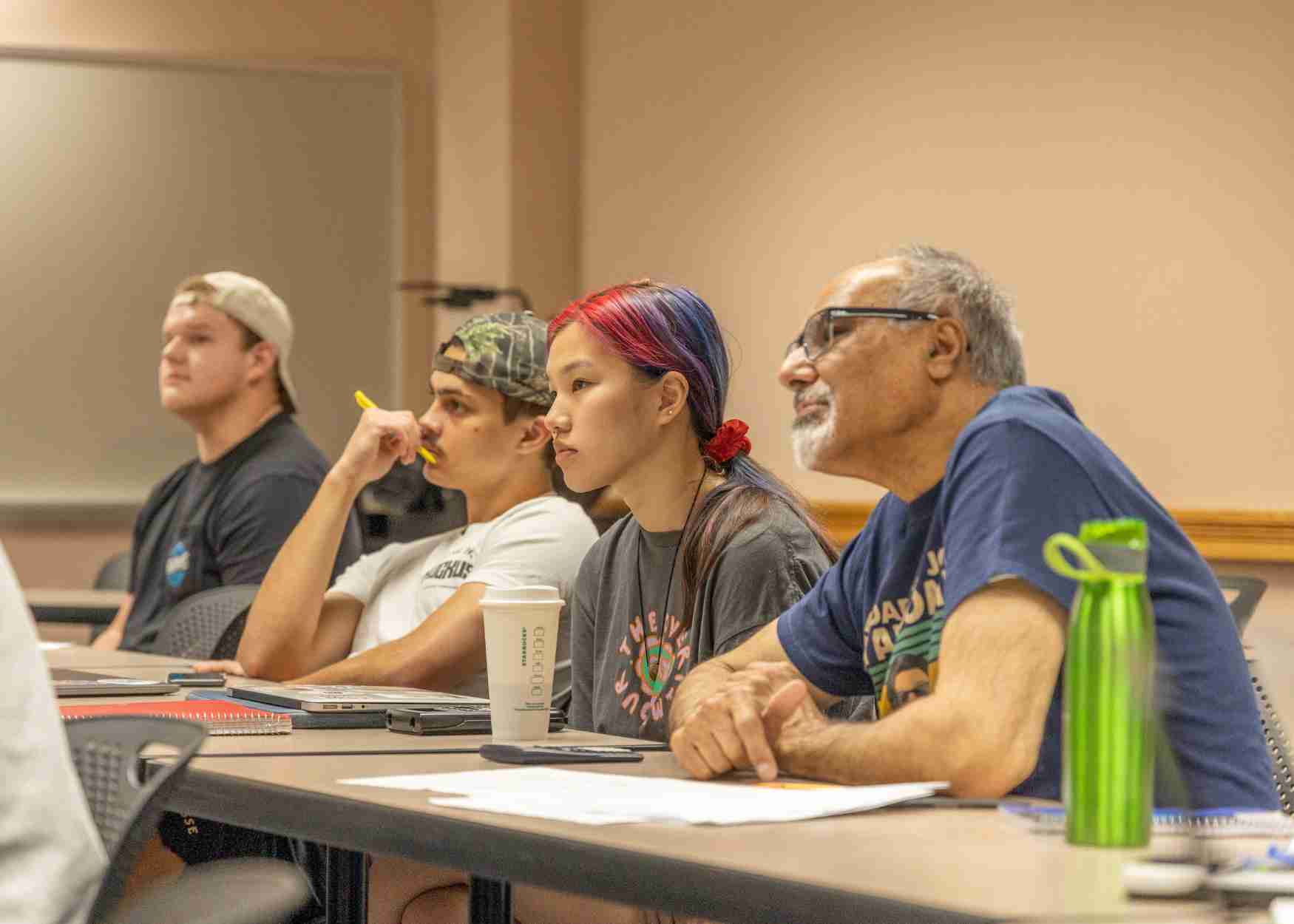 Professor sits with students at desk while listening to presentation