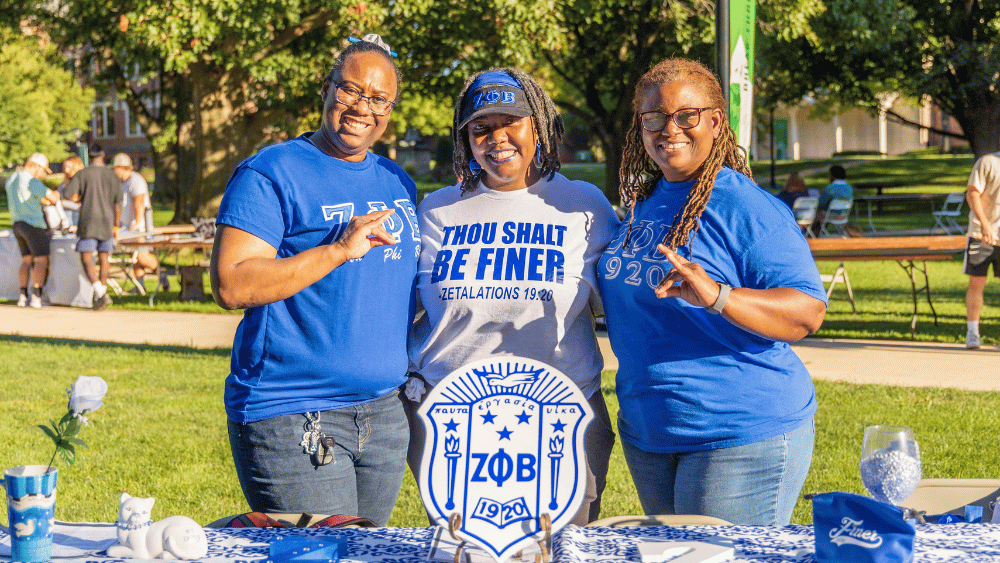 Three women from the local Zeta Phi Beta chapter stand at a table on the IWU quad during an RSO fair