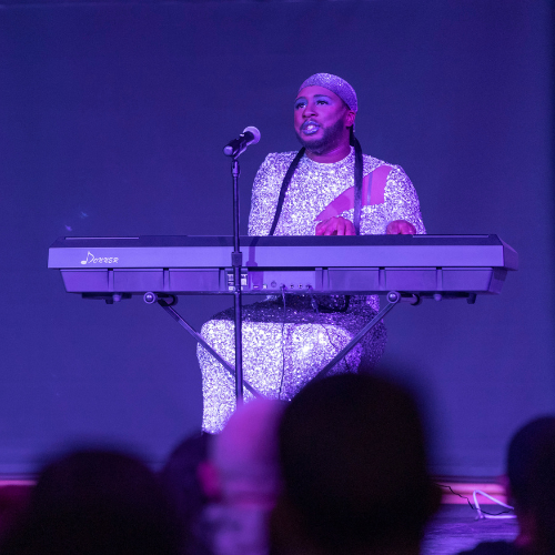 Reggie Cooke plays piano on stage in a silver sparkly dress as drag performer Reggay Boots