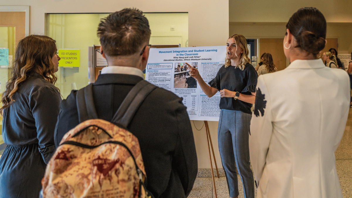 Three people stand listening to a student presenting research on a poster in IWU's State Farm Hall