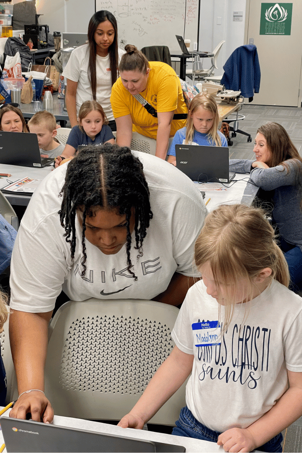 IWU students lean over tables to help children with 3d printing software on laptops