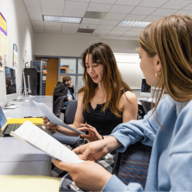Students talk in class holding papers and facing laptops