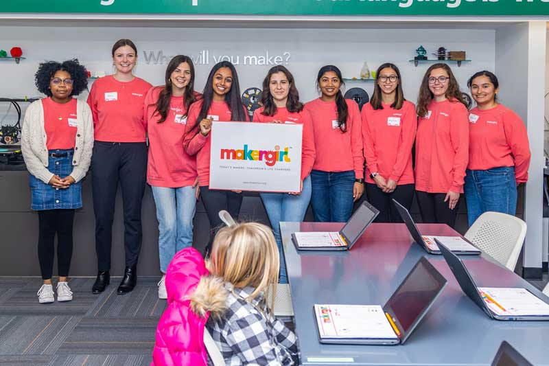 Nine IWU students look to the camera, smiling, and holding a MakerGirl sign at the first workshop for girls in the community in 2022.