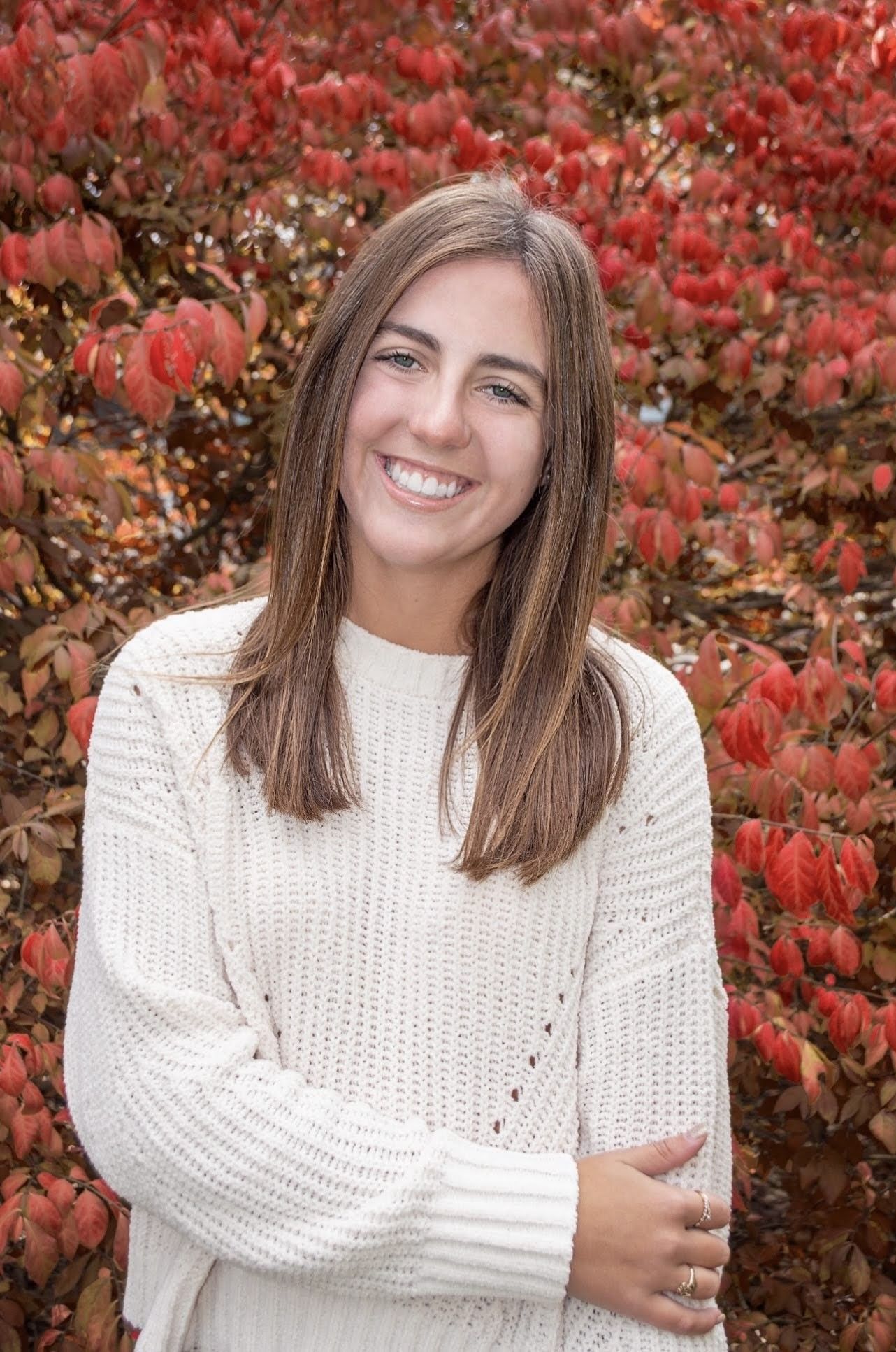 Female student in white sweater smiling in front of fall foliage