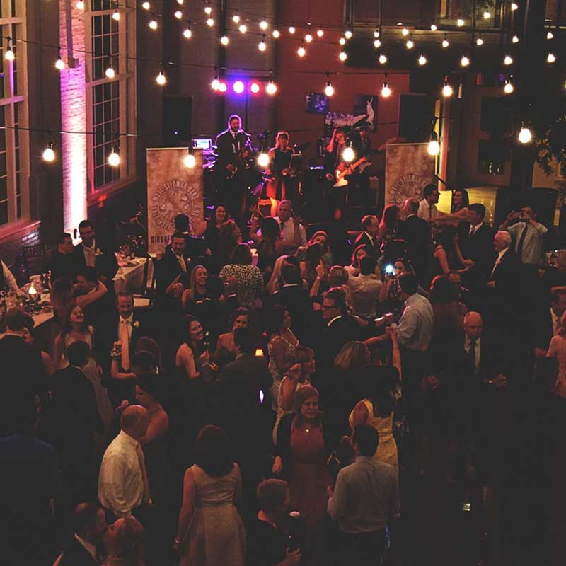A live band performs in the corner and people mingle on the dance floor, underneath edison light bulbs