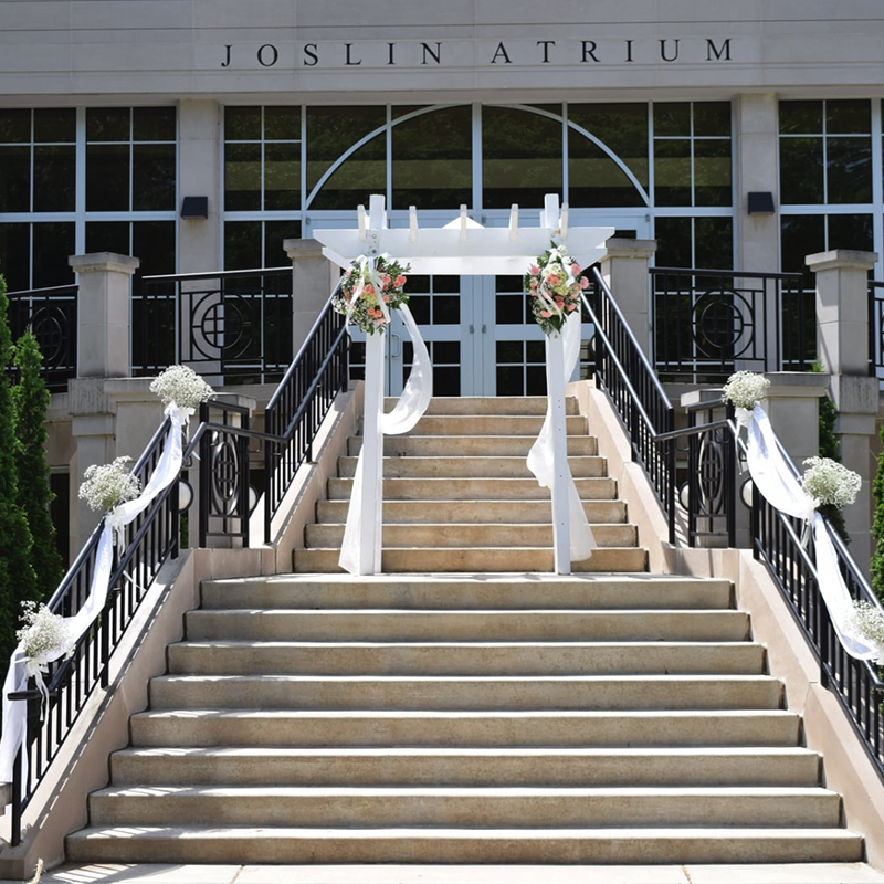 An arbor adorned with fabric and f lowers sits on the steps leading up to Joslin Atrium. Fabric and flowers are wrapped around the handrails.
